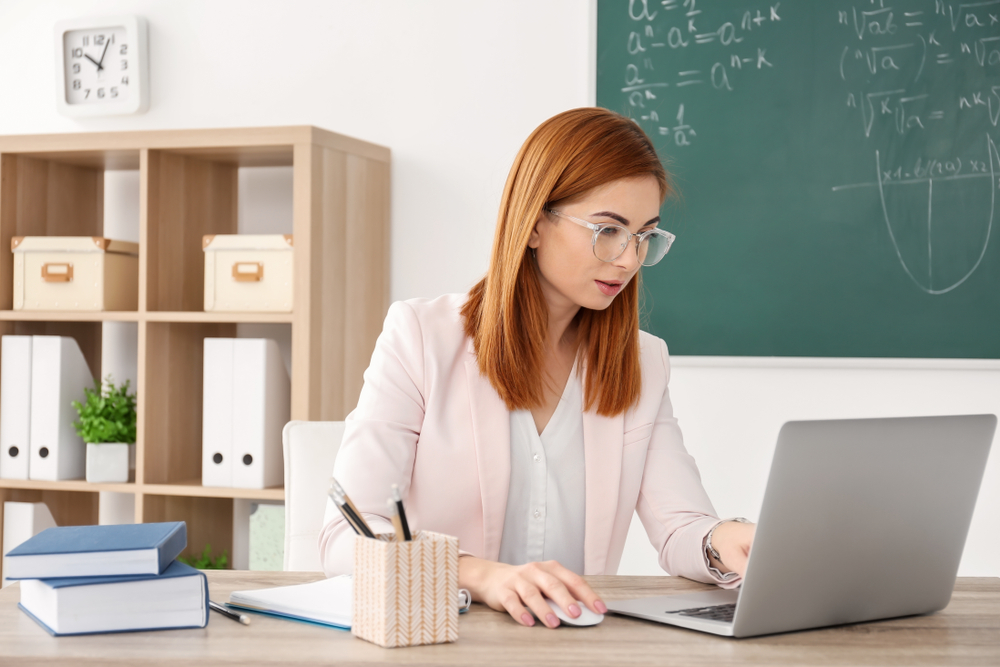 Beautiful,Young,Teacher,With,Laptop,Sitting,At,Table,In,Classroom